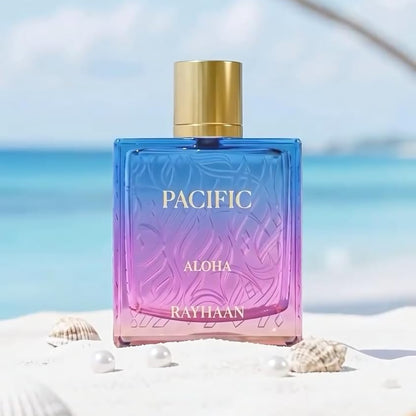 Rayhaan Pacific Aloha perfume bottle stands on white sand with shells and the ocean and palm tree branches blurred in the background. The bottle has a blue-to-purple gradient body with a gold cap.